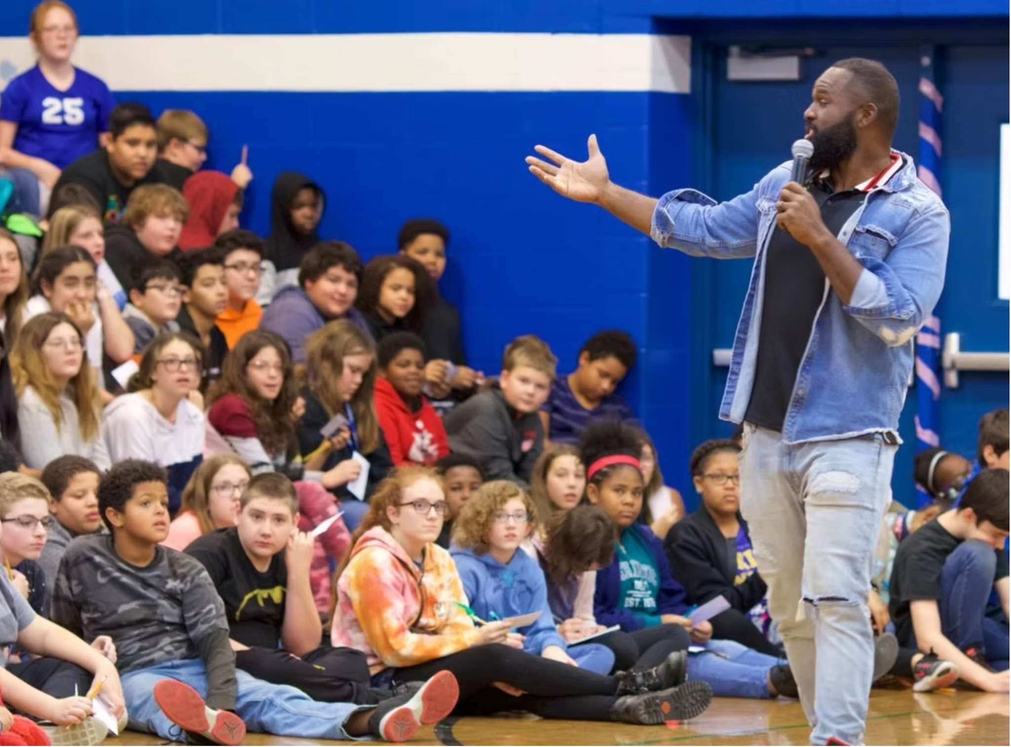 Andre Fluellen gesturing toward students seated on a school gym floor while speaking into a microphone